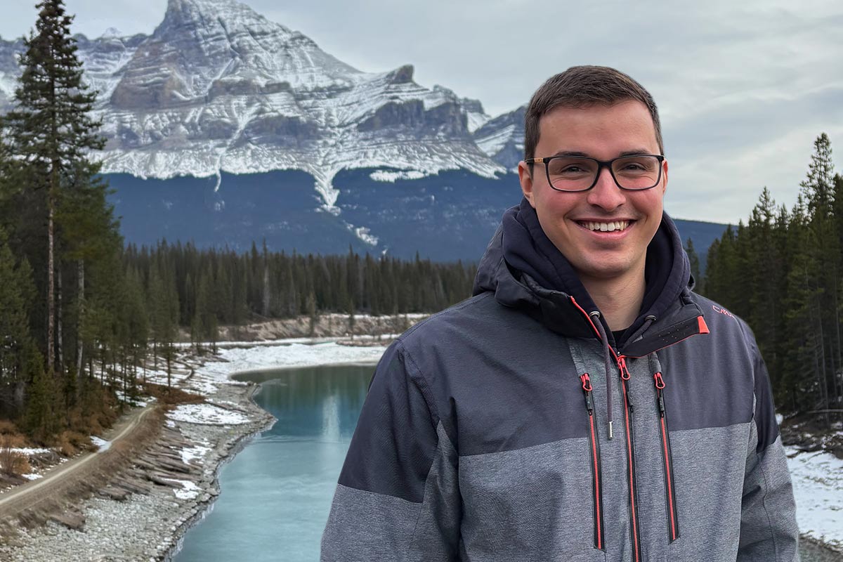 Junger Mann mit Brille und grauer Jacke lächelt vor verschneiter Berglandschaft mit Fluss und Tannenwald.